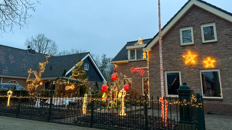 Twee huizen versierd met kerstverlichting, inclusief sterren en figuren, achter een hek met lichtjes.