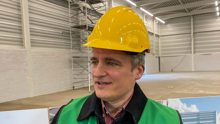 Man met een gele bouwhelm en groene veiligheidshes op een bouwlocatie met steiger op de achtergrond.