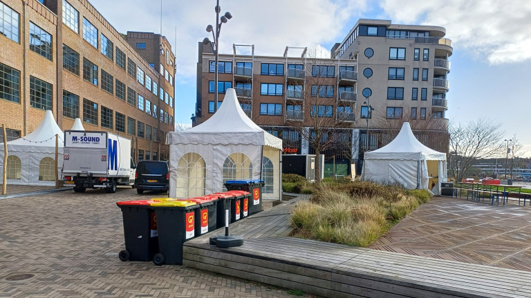 Pleintje met tenten, een vrachtwagen en vuilnisbakken voor een bakstenen gebouw en moderne appartementen.