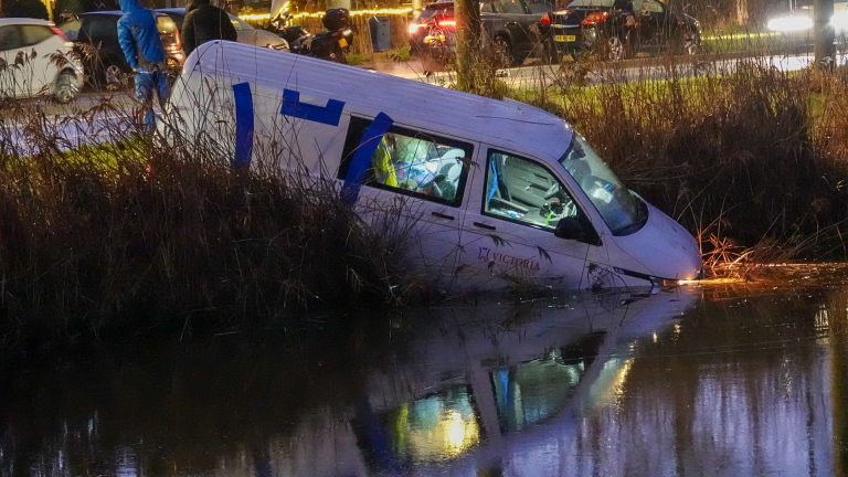 Een bestelwagen half ondergedompeld in een sloot, omringd door riet, terwijl mensen op de weg staan te kijken.
