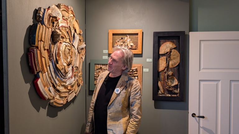 Een man kijkt naar een kunstwerk aan de muur gemaakt van boeken en papier, met andere kunstwerken op de achtergrond in een galerij.