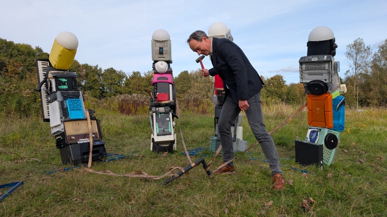 Een man in een jasje gebruikt een hamer om een paal in de grond te slaan naast kunstwerken gemaakt van gerecyclede elektronische apparaten op een grasveld.