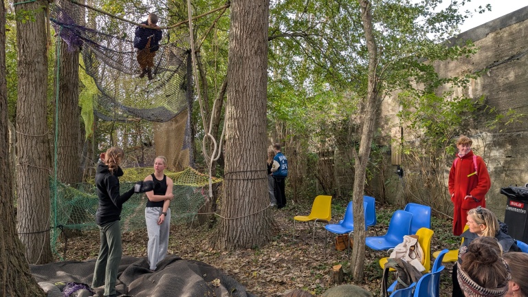 Een groep mensen in een bosrijke omgeving met netten tussen de bomen waar iemand op klimt. Er zijn kleurrijke stoelen, een persoon in een rode jas en anderen die toekijken of praten. De omgeving lijkt een speelse en creatieve ruimte.