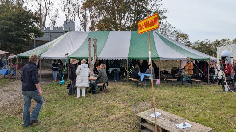 Een groen-wit gestreepte tent met mensen ervoor en erin tijdens een evenement. Een geel-rood bord met de tekst 'BOUWENWALKENNAAR' staat in de voorgrond. In de tent zitten en staan mensen, en er is een persoon met machines aan de rechterkant. Op de achtergrond staan bomen en een kasteelachtig gebouw.
