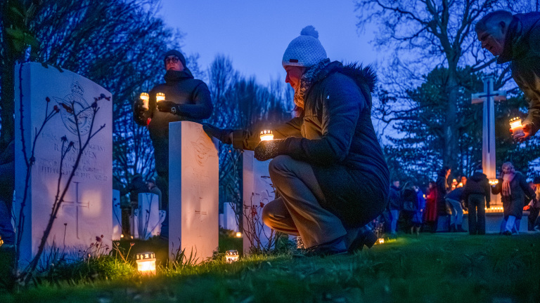 Mensen plaatsen kaarsen bij grafstenen op een begraafplaats bij avondschemering, omringd door bomen en andere aanwezigen.