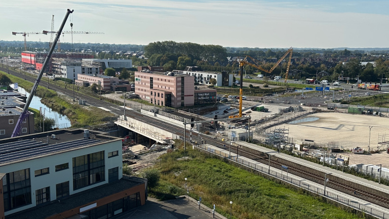 Bouwterrein met hijskranen nabij een spoorlijn en gebouwen onder constructie, omringd door groen en wegen.