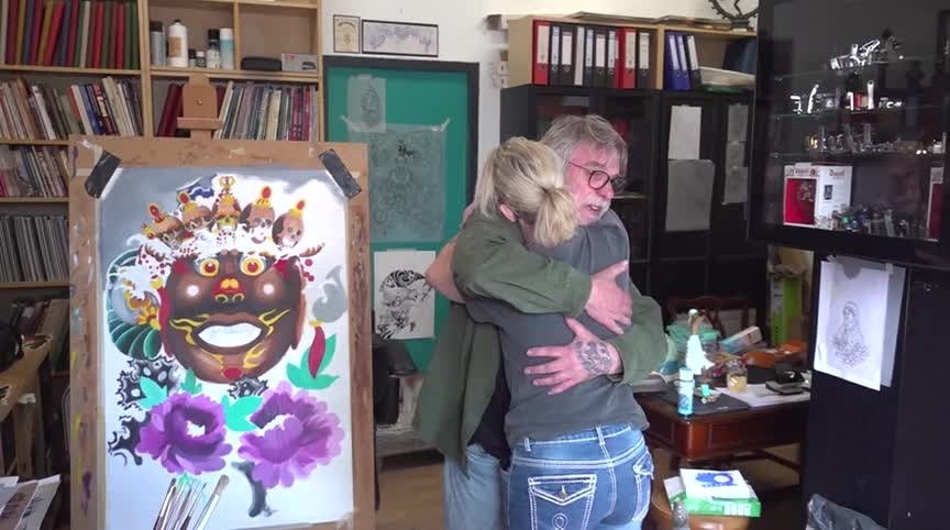 Twee mensen omhelzen elkaar in een kunststudio, met kleurrijke schilderijen en boeken op de achtergrond.