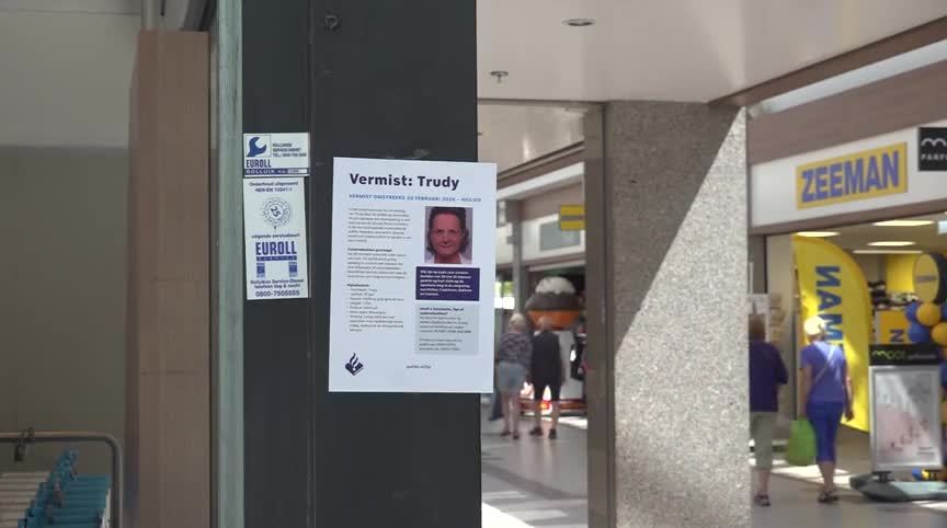 Opsporingsposter voor een vermist persoon op een paal in een winkelcentrum, met voorbijgangers en een Zeeman-winkel op de achtergrond.