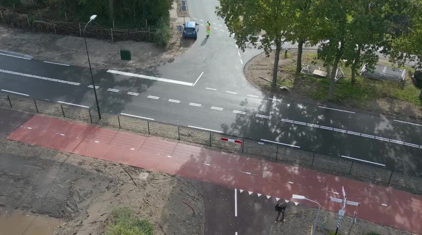 Luchtfoto van kruising met rood fietspad langs de weg, bomen en een auto met twee personen in fluorescerende kleding