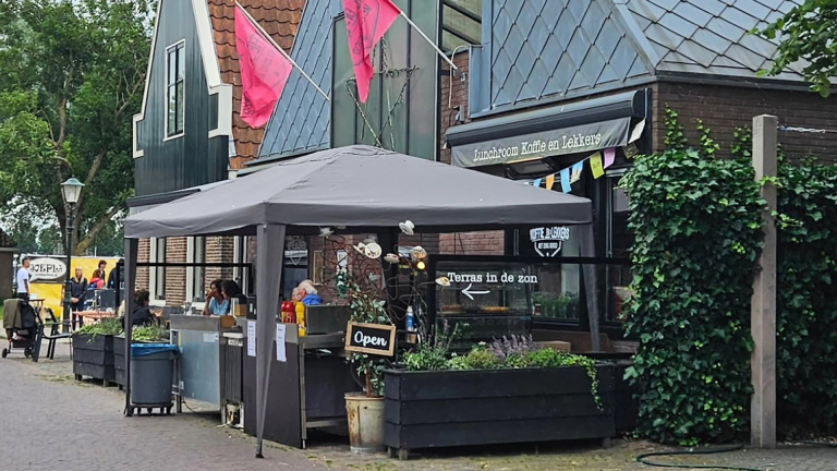 Terras van lunchroom met grote parasol en bordje Open, bij gevel met tekst Lunchroom Koffie en Lekkens