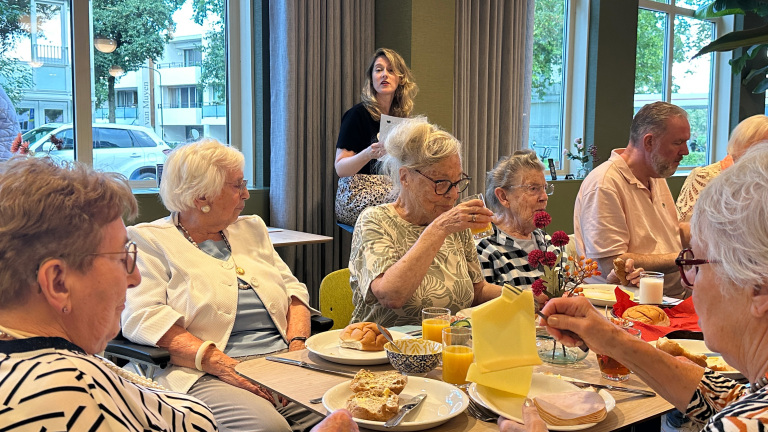 Groep ouderen zit aan een gedekte tafel met broodjes en sap, terwijl een vrouw staand iets toespreekt