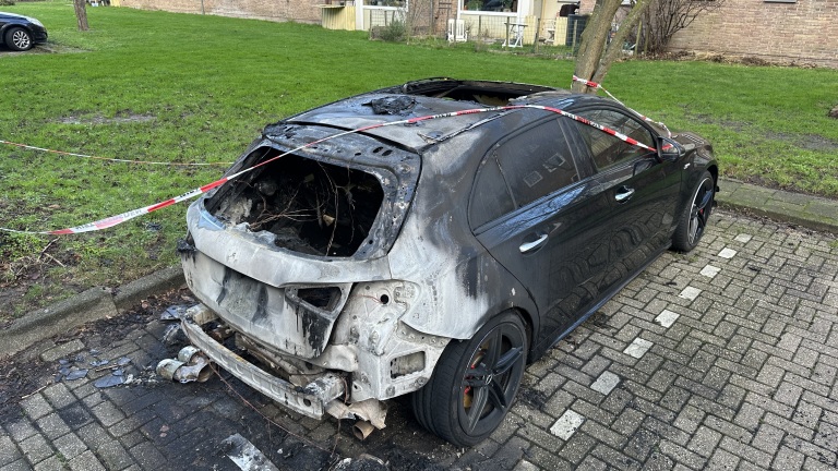 Uitgebrande zwarte auto met brandschade aan de achterkant, afgebakend met politielint, geparkeerd op een stenen parkeerplaats.