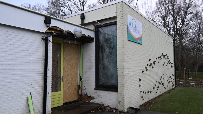 Wit gebouw met beschadigde deur en gebroken raam, naast een klimmuur en bord met opschrift "Wild Formid".