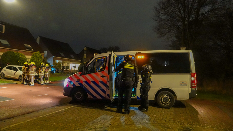 Politieagenten en brandweerlieden bij een politievoertuig en huizen in een woonwijk, 's avonds.
