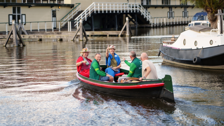 Vijf mensen in een sloep op een kanaal, zwaaiend naar de camera, met boten en een houten gebouw op de achtergrond