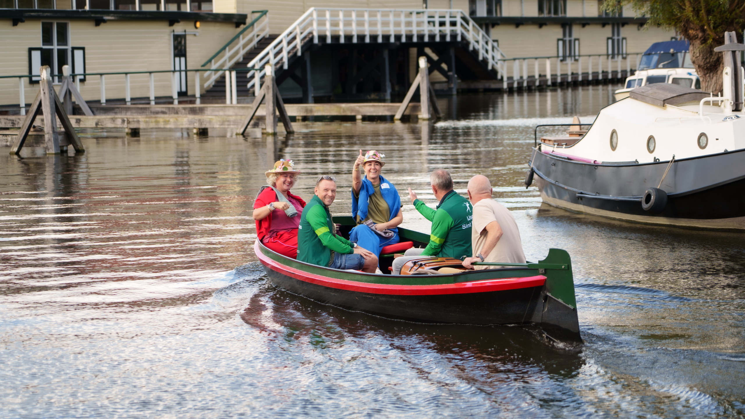 Vijf mensen in een sloep op een kanaal, zwaaiend naar de camera, met boten en een houten gebouw op de achtergrond