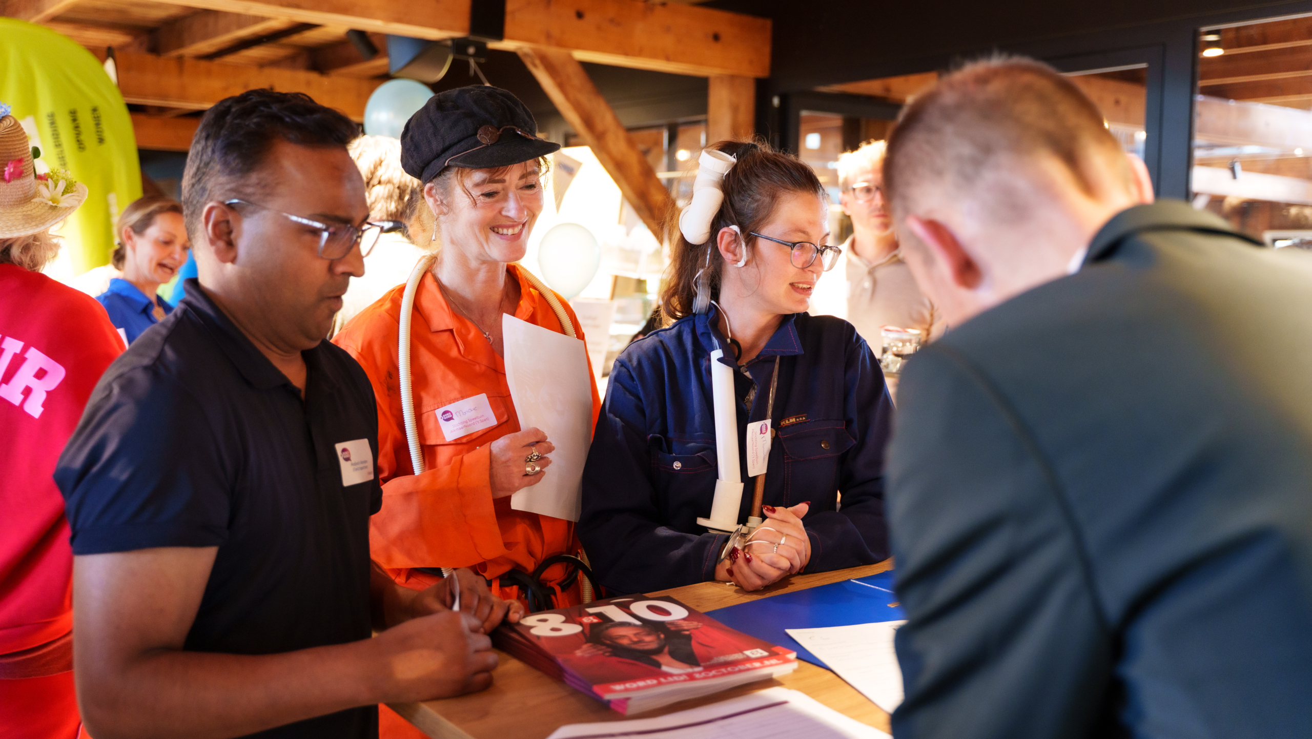 Drie deelnemers in werkoveralls staan aan een balie en overleggen met een medewerker tijdens een evenement
