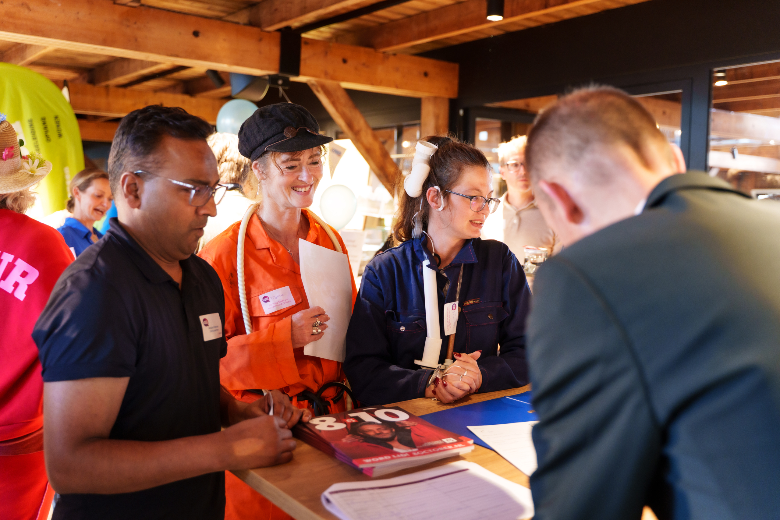 Drie deelnemers in werkoveralls staan aan een balie en overleggen met een medewerker tijdens een evenement