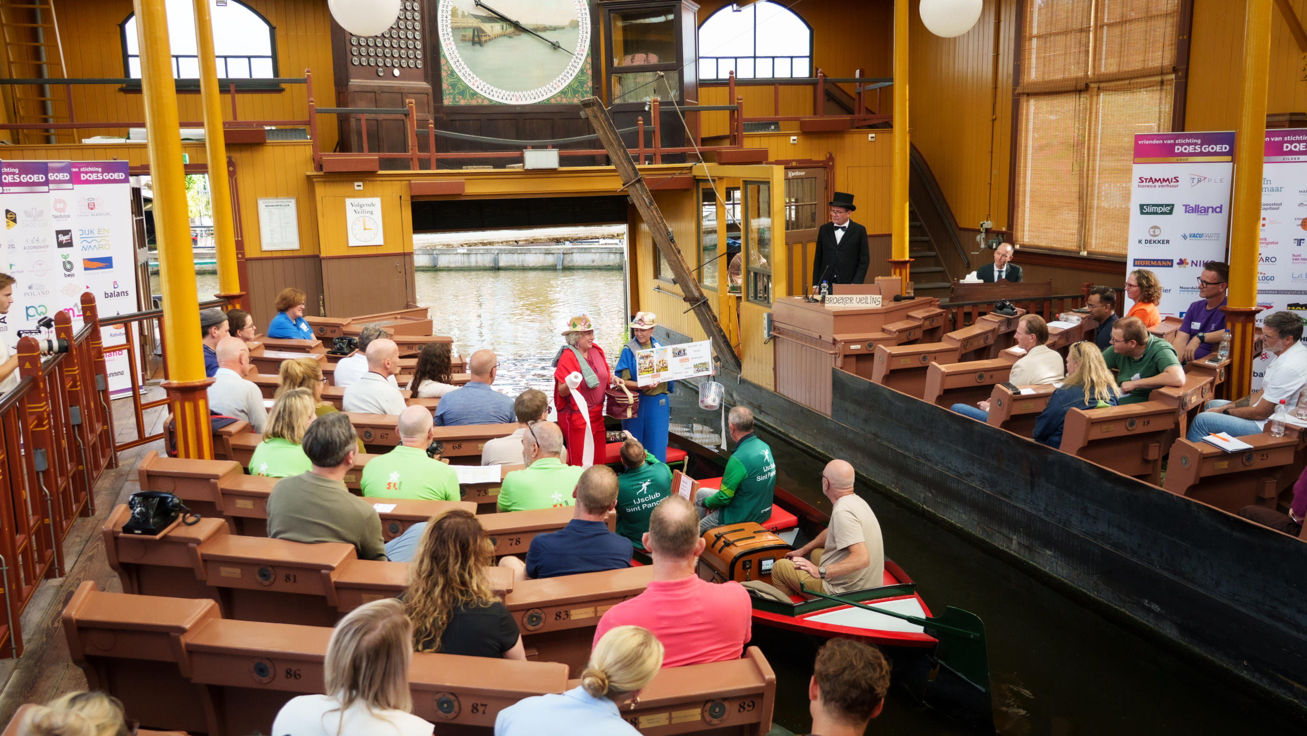 Publiek op houten banken bij een veilingzaal aan het water, met veilingmeester en twee vrouwen op een boot in het midden