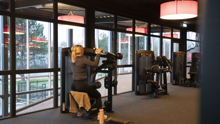 Vrouw traint op een krachtapparaat in een sportschool met ramen en rode lampen.