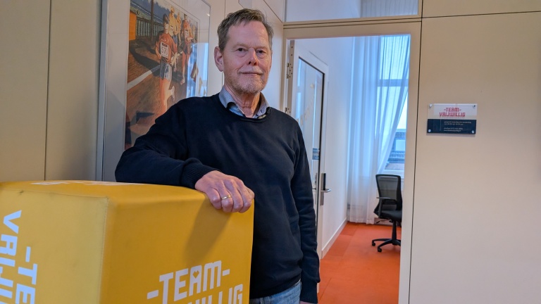 Man leunt tegen een gele doos met de tekst "TEAM-VRIJWILLIG" in een kantoor met een oranje vloer.