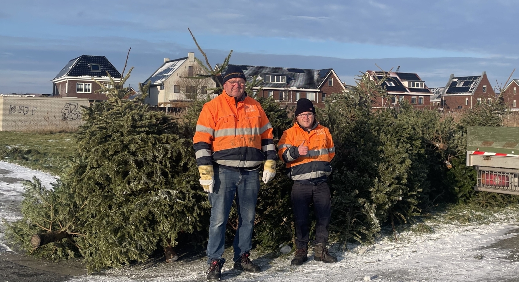 Twee mannen in oranje jassen staan voor een stapel kerstbomen naast een vrachtwagen in een woonwijk.