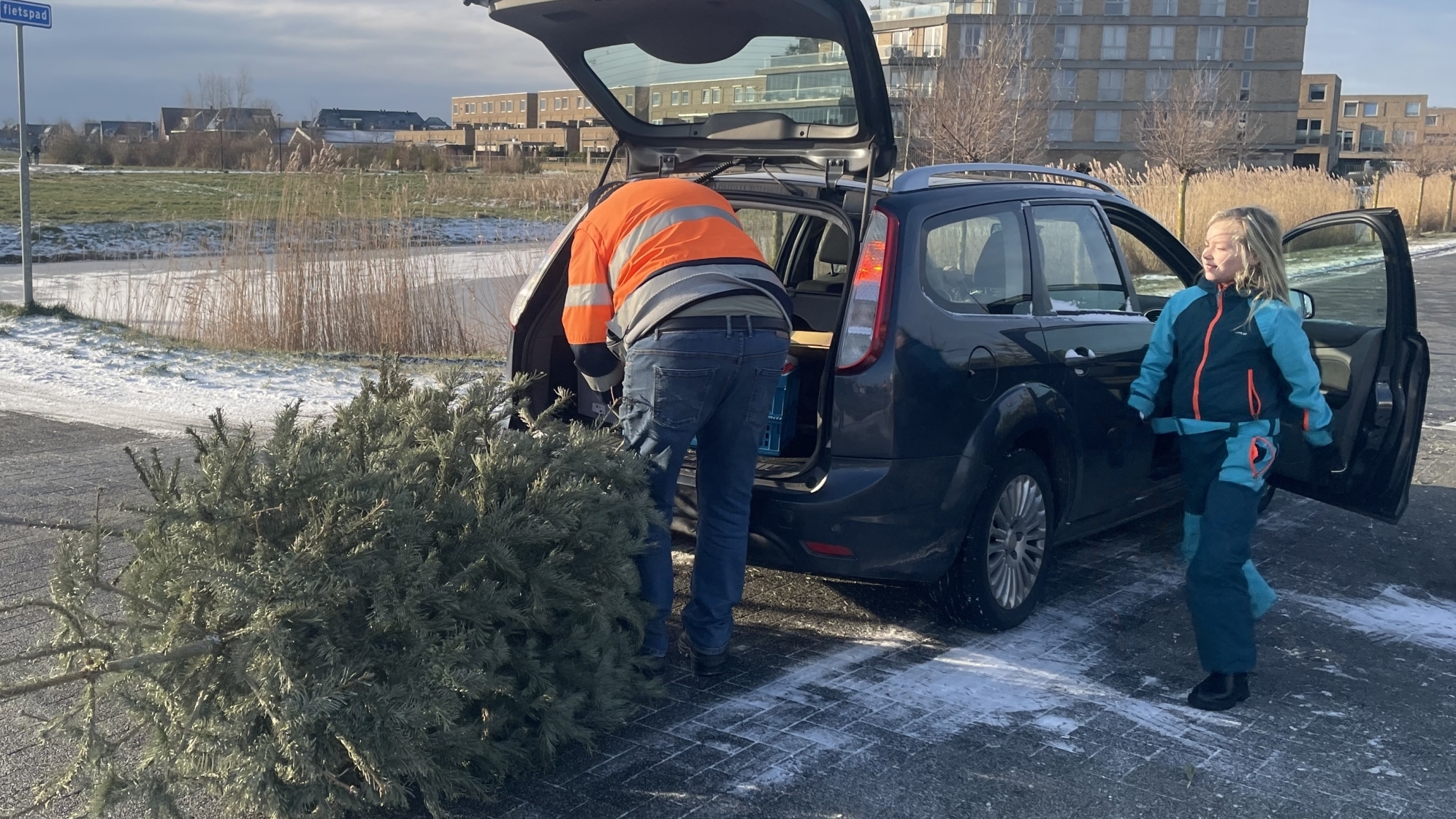 Een persoon in een oranje jas laadt een kerstboom in de kofferbak van een auto, terwijl een kind ernaast staat.