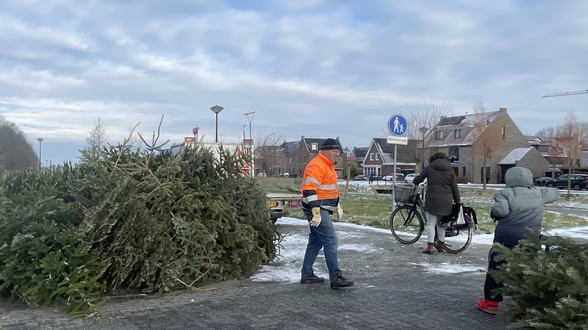 Stapels kerstbomen langs een besneeuwde weg met een persoon in feloranje jas die voorbijloopt, en een fietser met kind.