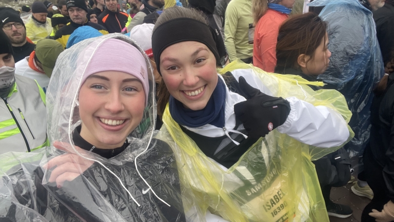 Twee lachende vrouwen in regenponcho's klaar voor een evenement, omringd door andere deelnemers in warme kleding.