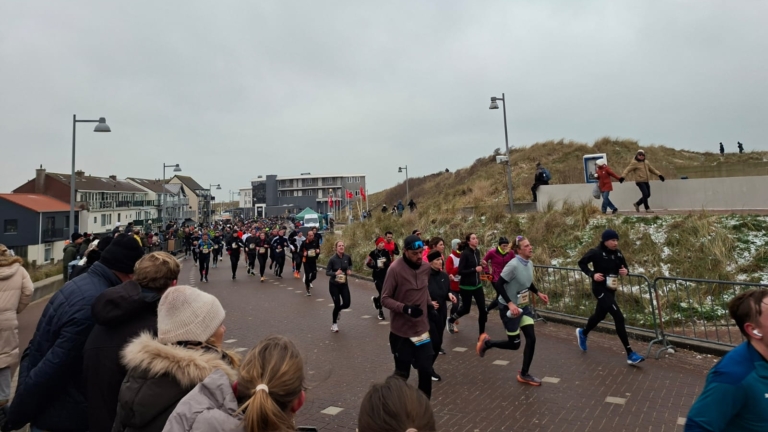 Een groep hardlopers op een besneeuwd pad langs duinen, omringd door toeschouwers en gebouwen op de achtergrond.