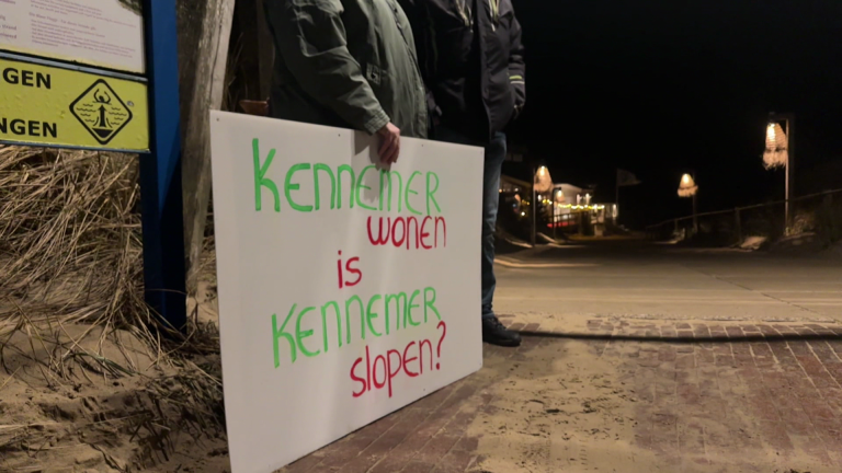 Twee mensen houden een bord met de tekst "Kennemer wonen is Kennemer slopen?" naast een verlicht pad in de nacht.