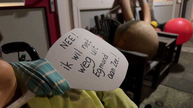 Papieren bordje met de tekst "NEE! ik wil niet weg uit Egmond aan Zee!" bevestigd aan een rolstoel in een kamer.