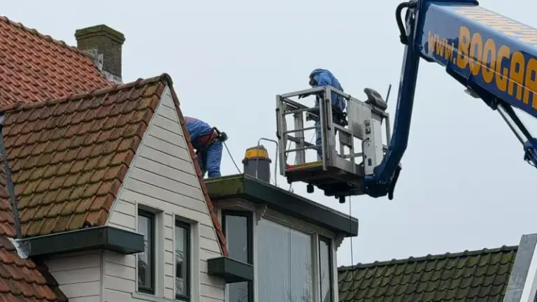 Twee arbeiders in veiligheidsharnassen werken vanuit een hoogwerker aan de schoorsteen van een huis met pannendak.