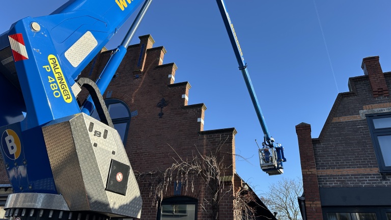 Een hoogwerker van het merk Palfinger wordt gebruikt bij een bakstenen huis met trapgevel onder een blauwe hemel.