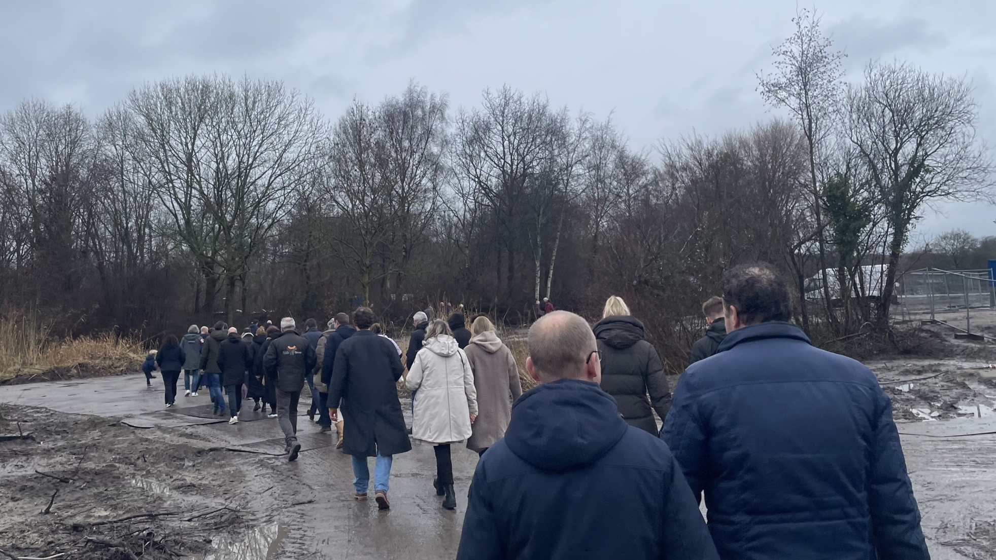 Een groep mensen loopt over een modderig pad langs bomen en een bouwterrein op een bewolkte dag.