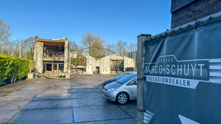 Afgebrand garagegebouw met naambord "Marco Schuyt Occasionsdealer" op de voorgrond en geparkeerde auto's.