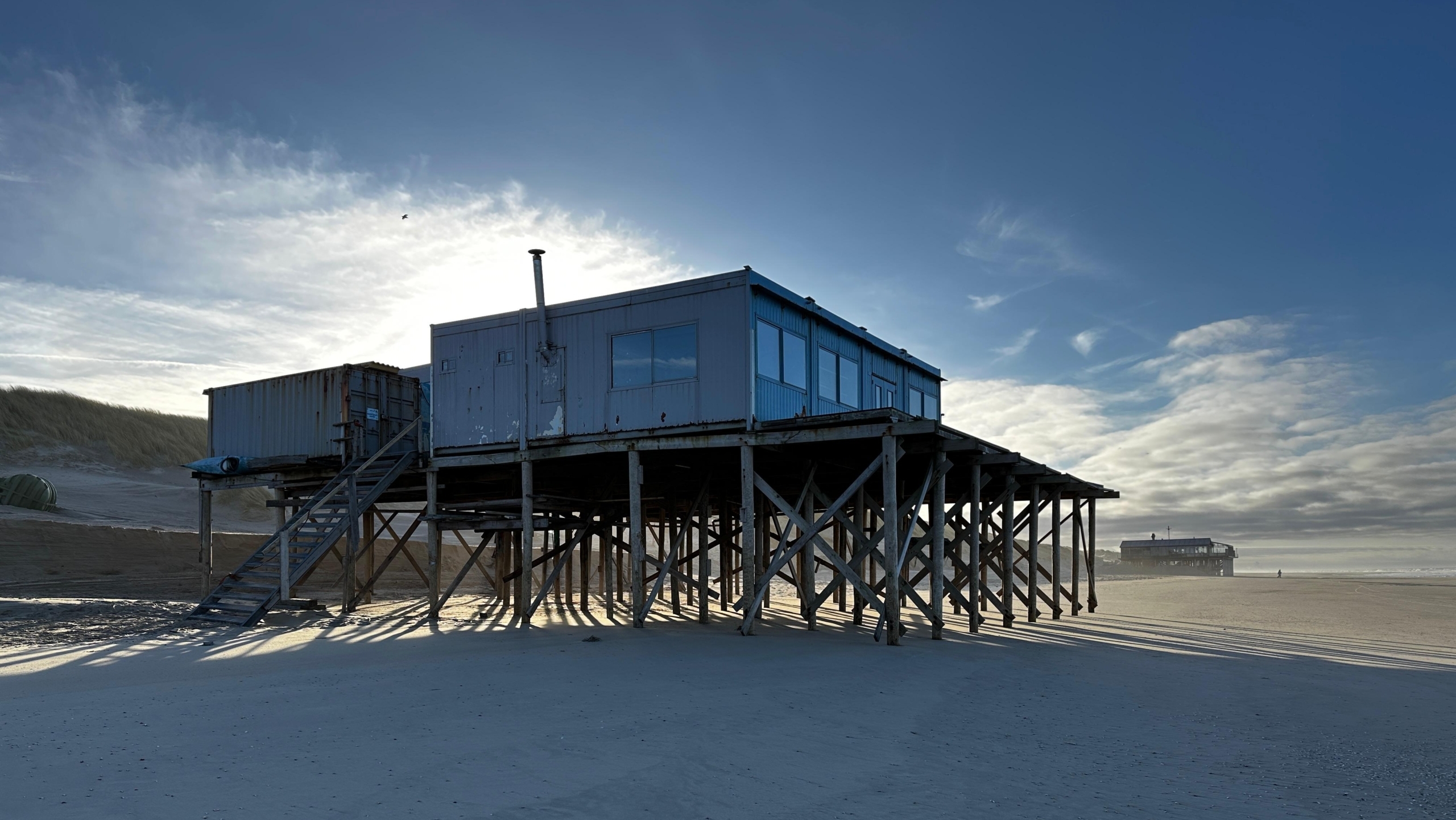 Een strandpaviljoen op stelten bij zonsondergang met een uitgestrekt zandstrand en duinen op de achtergrond.