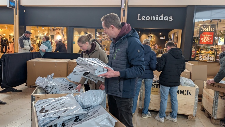 Mensen bladeren door dozen met plastic verpakte producten in een winkel met een Leonidas winkel op de achtergrond.