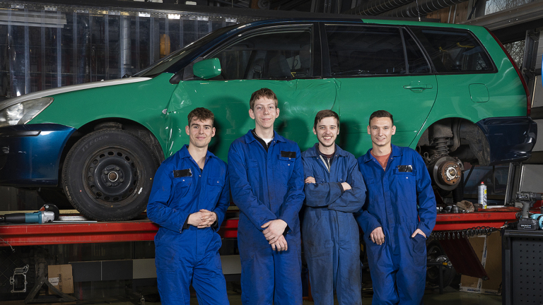 Vier monteurs in blauwe overalls poseren voor een auto op een hefbrug in een werkplaats.
