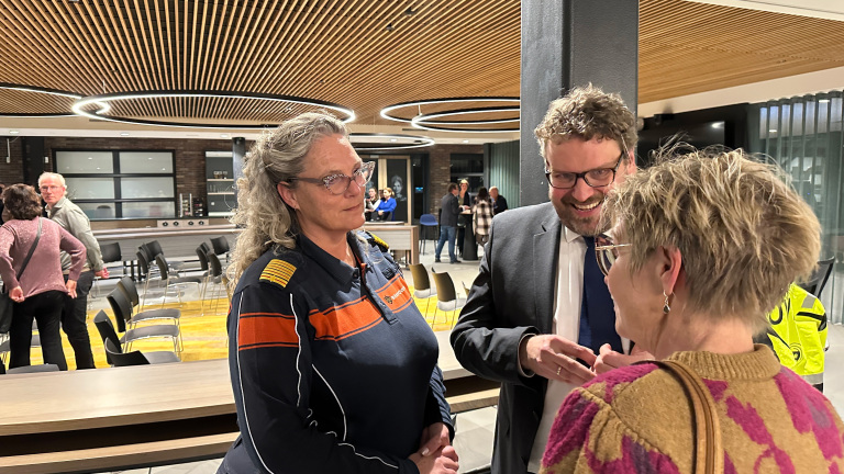 Drie mensen in gesprek in een moderne hal met houten plafond en stoelen; één draagt een uniform met oranje en navy strepen.