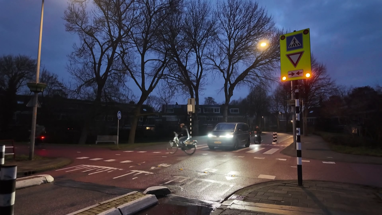 Een fietser steekt 's avonds een weg over terwijl een auto wacht bij een verlichte kruising met verkeersborden en bomen.