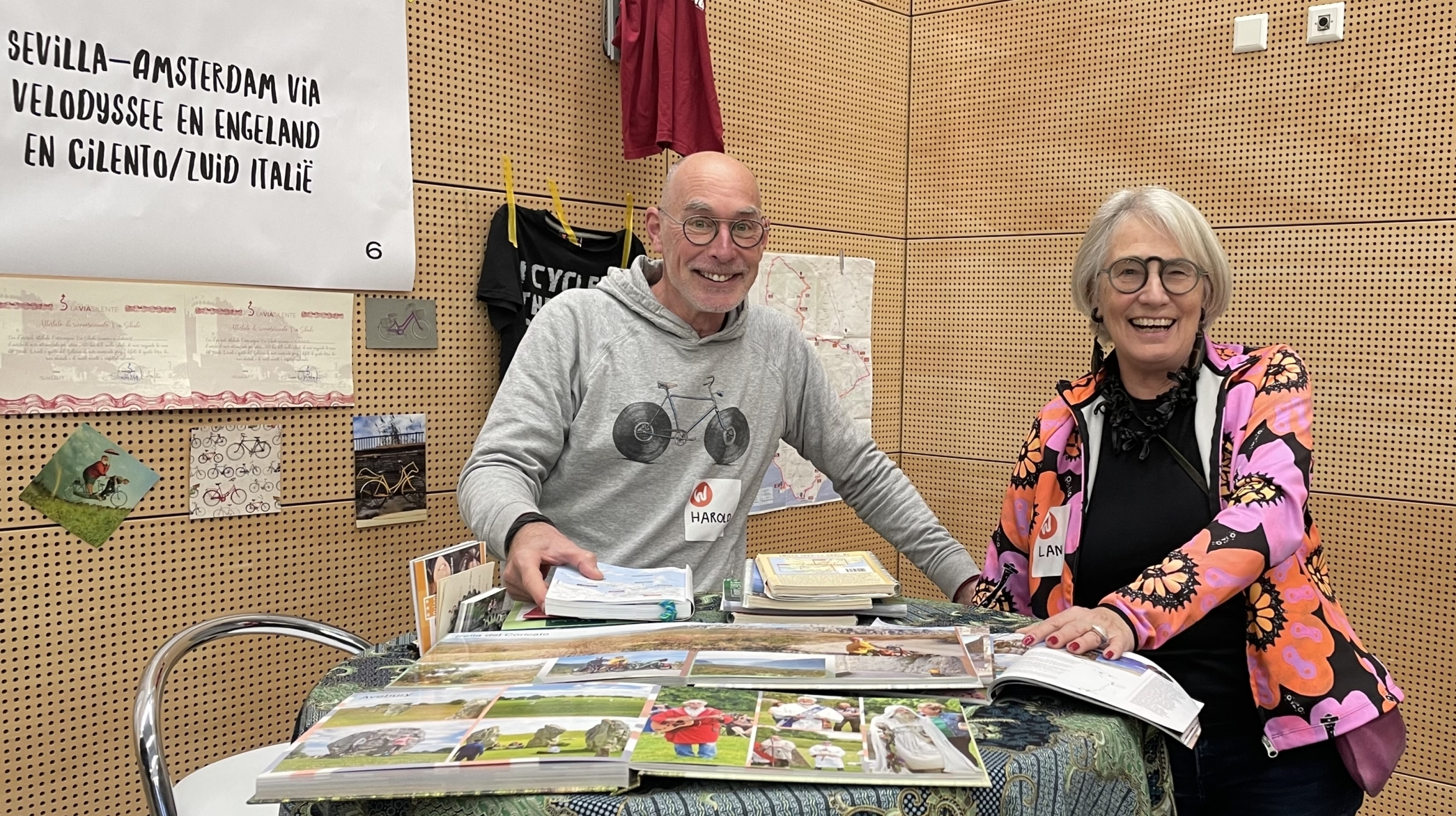 Twee mensen glimlachen achter een tafel vol reisfolders en kaarten; een poster met een routebeschrijving hangt aan de muur.