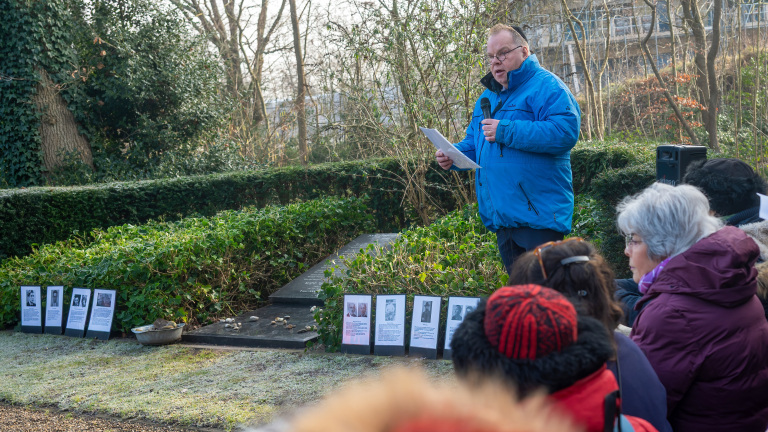 Man in een blauwe jas spreekt voor een groep mensen bij een gedenkplaats omringd door struiken en fotoborden.