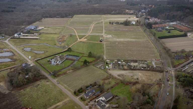Luchtfoto van een landelijke omgeving met weilanden, enkele huizen en omliggende bossen, wegen en percelen.