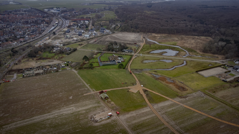 Luchtfoto van een landschap met weilanden, bos, enkele huizen en een weg met voertuigen en bouwactiviteiten bij een stad.
