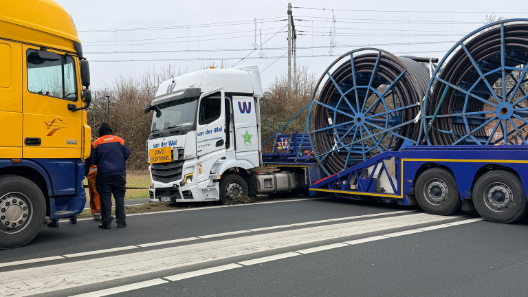 Twee vrachtwagens, een gele en een witte, staan op de weg met een grote haspel op een trailer; personen ernaast.