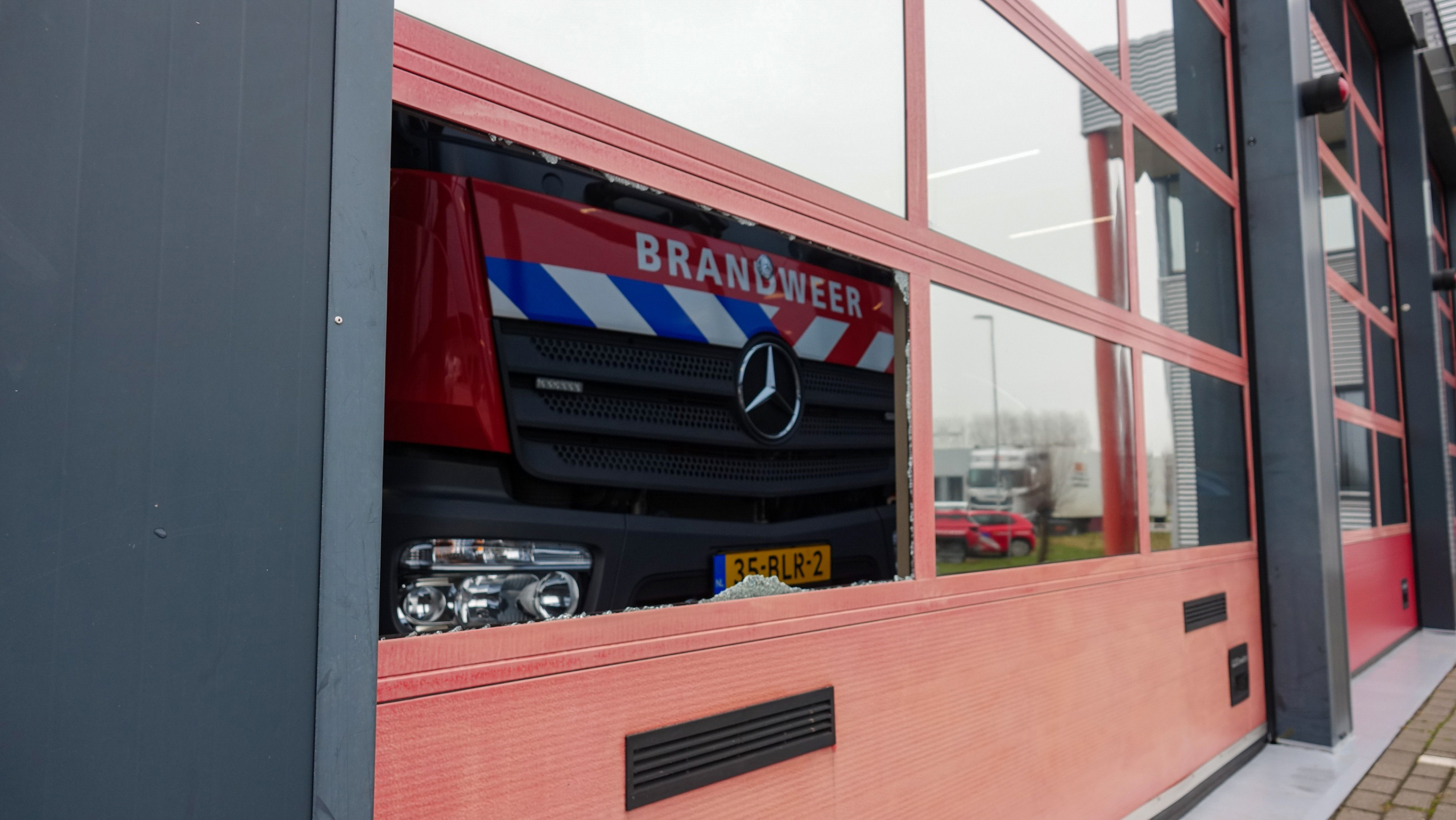 Rode brandweerwagen achter de glazen deur van een brandweerkazerne met het Mercedes-Benz logo en kenteken zichtbaar.