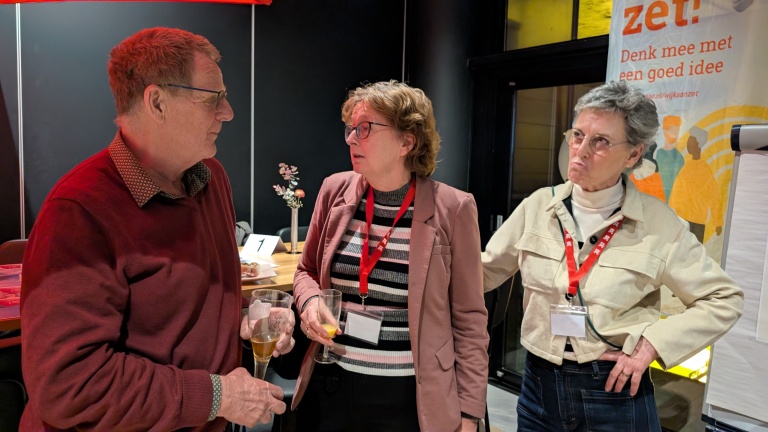 Drie mensen in gesprek bij een tafel met glazen; op de achtergrond een bord met de tekst "Denk mee met een goed idee".