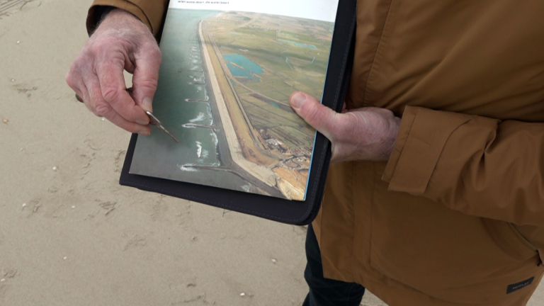 Een persoon in een bruine jas wijst met een pen naar een foto van een kustlandschap op een strand.
