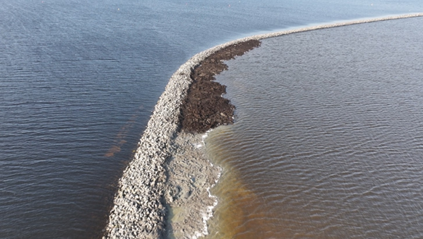 Een luchtfoto van een dijk die water aan beide zijden scheidt, met een donkere strook vuil op de rechterkant.